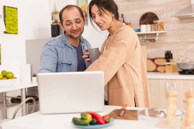 A couple smiling and holding hands while looking at a laptop displaying a balanced budget. The setting is a modern kitchen with natural light.