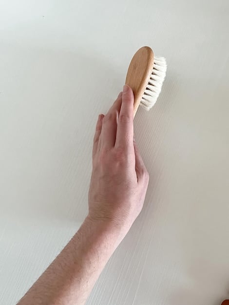 Close-up shot of a person using a scrub brush to clean the surface of a wooden deck. The focus is on the scrubbing action, showing the bristles of the brush removing dirt and grime from the wood grain. The surrounding area is wet, indicating recent cleaning.