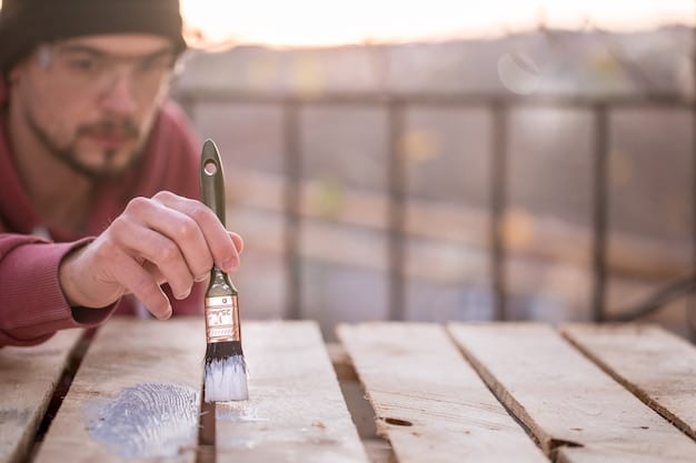 Medium shot of a person applying deck sealant to a wooden deck using a brush. The brush is loaded with sealant, and the person is carefully applying it to the wood grain. The surrounding area is already sealed, showing a slightly darker tint.