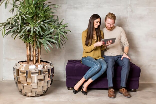 A couple sitting on a couch, surrounded by plants and sunlight, looking at a laptop with a financial forecast chart on the screen. The scene conveys a sense of peace and future planning.